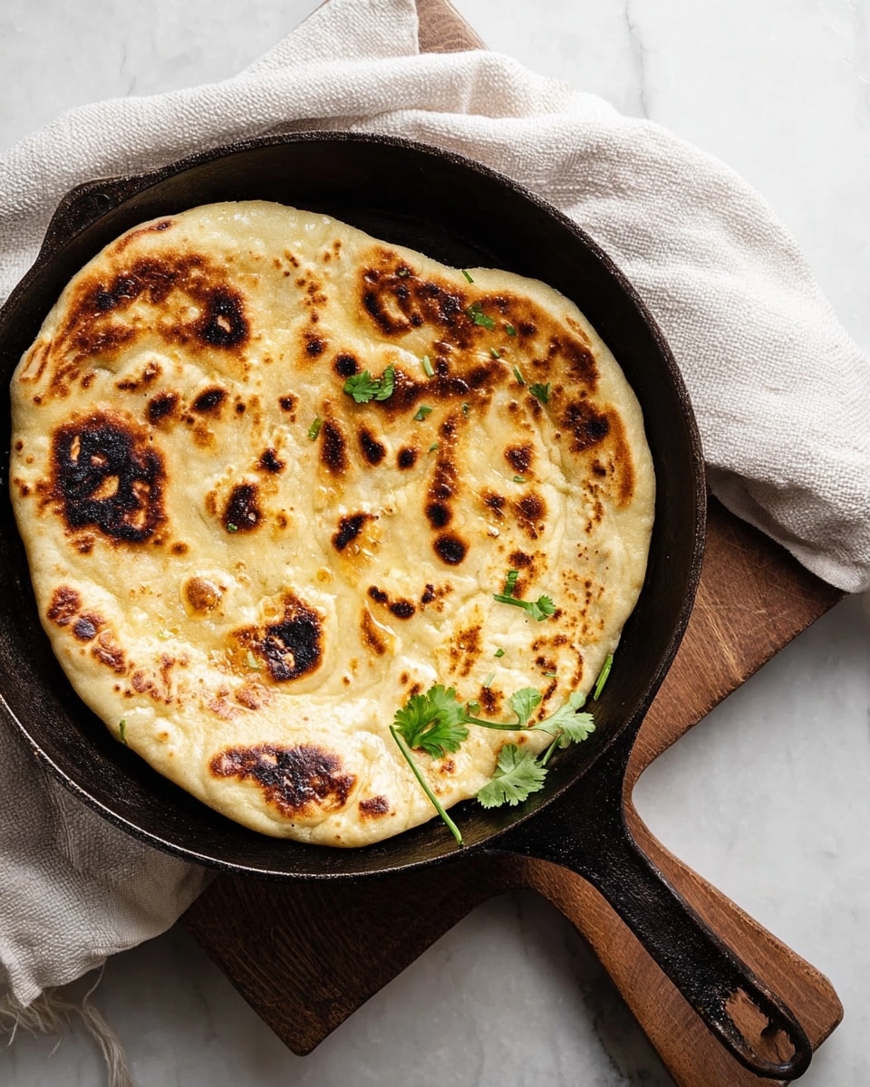 A round, flat naan bread with golden brown and dark charred spots sits inside a black cast iron skillet placed on a small wooden board over a white marbled surface. The naan has a slightly puffy texture with uneven bubbles and is garnished with a few small green cilantro leaves on one side. A white cloth is partly visible beside the skillet, adding a soft contrast. photo taken with an iphone --ar 4:5 --v 7