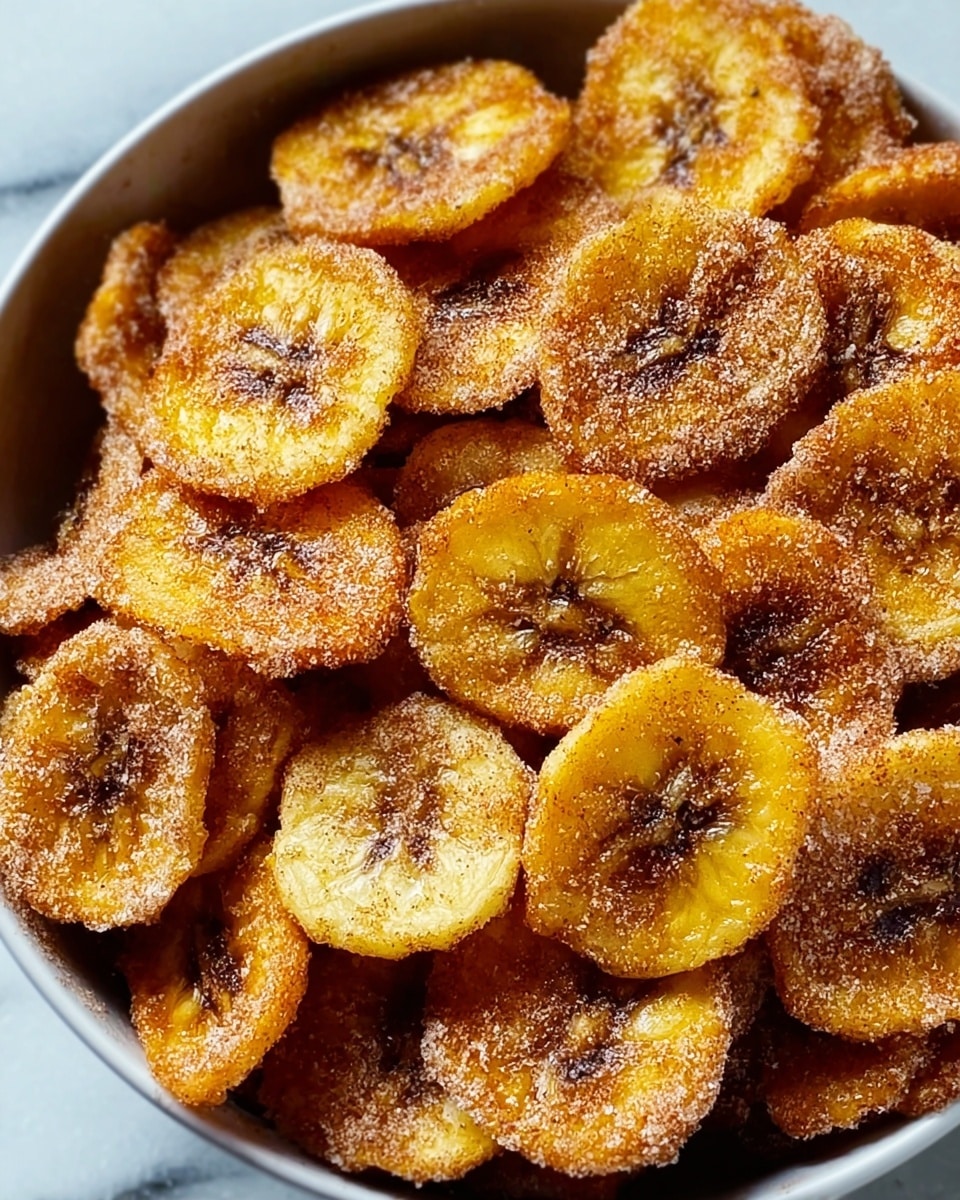 A black frying pan lined with white paper is filled with many round, golden-yellow plantain chips, each chip coated with a layer of light brown cinnamon sugar, giving a grainy texture on top. The chips show some darker brown spots where they are fried more, making the edges look crispy and slightly curled. The pan is placed on a white marbled surface. Photo taken with an iphone --ar 4:5 --v 7