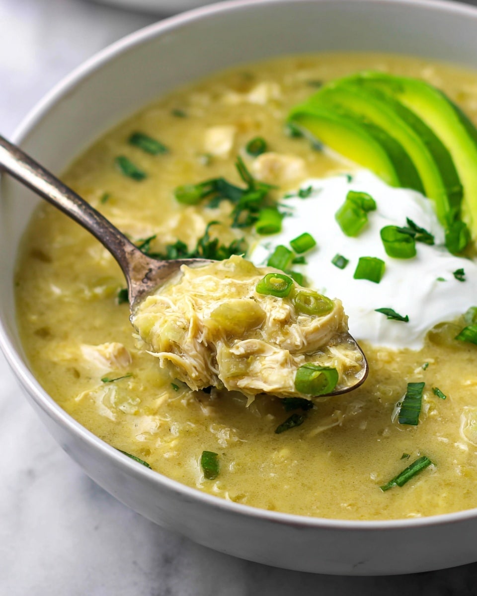 A white bowl is filled with creamy, chunky soup that has a light yellow-green color, showing pieces of tender shredded chicken mixed in. On the surface, there is a dollop of smooth white sour cream placed near the top center, next to three slices of fresh green avocado arranged in a neat row on the right side. Bright green chopped scallions are scattered across the top, adding color and texture. A silver spoon in the bowl scoops up some of the thick soup, capturing a close-up of the mixture with visible herbs and chicken strands. The bowl sits on a white marbled surface. photo taken with an iphone --ar 4:5 --v 7