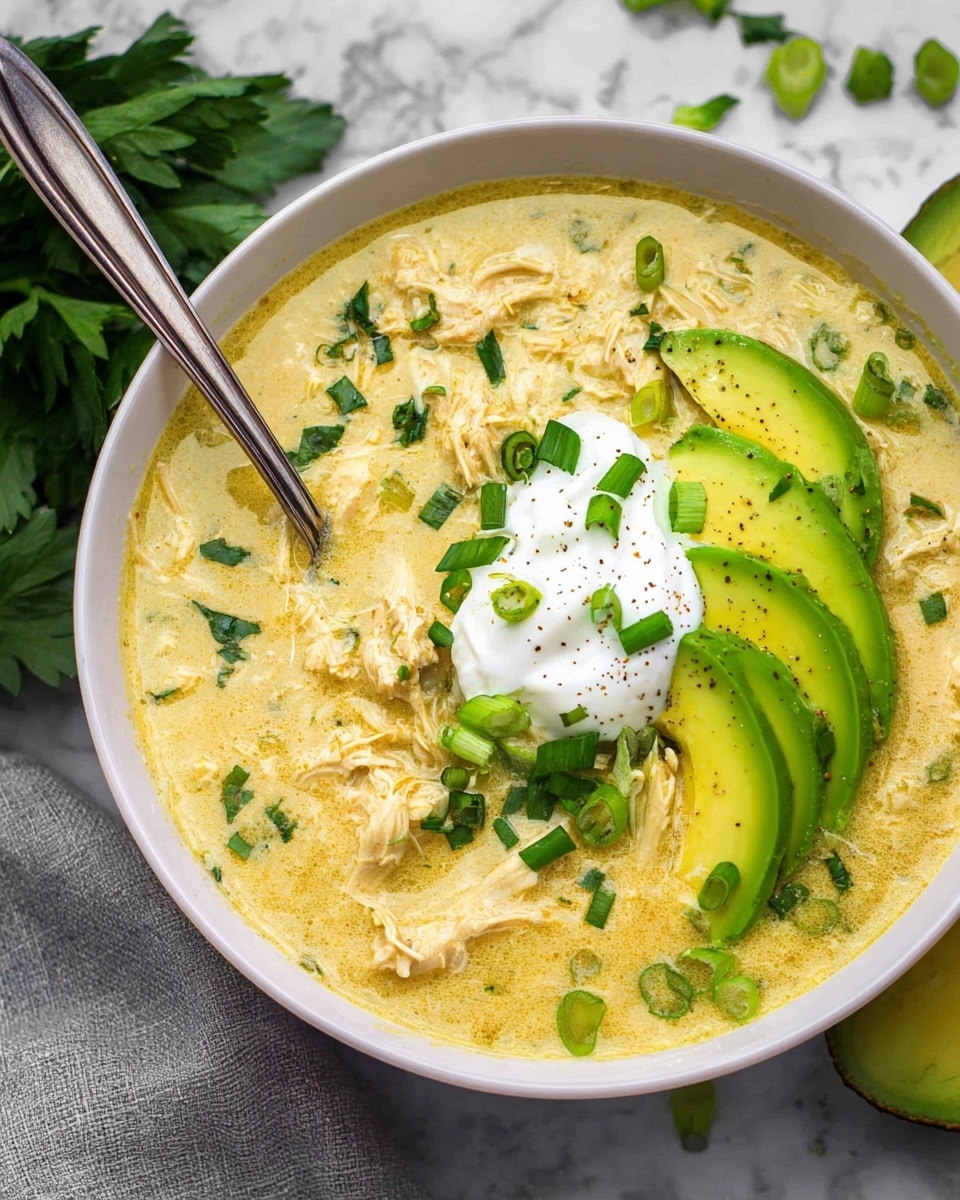A white bowl filled with a creamy, pale yellow soup that has a thick texture with visible small chunks of shredded chicken mixed in. On top, there is a dollop of white sour cream placed slightly off-center, beside thin slices of bright green avocado arranged in a small stack on the right side. Scattered across the surface are fresh chopped green onions and small bits of fresh green herbs, adding a pop of color and texture. A metallic spoon is partially dipped into the soup on the left side of the bowl. The bowl is placed on a white marbled surface with a few avocado slices and herb leaves scattered around it. Photo taken with an iphone --ar 4:5 --v 7