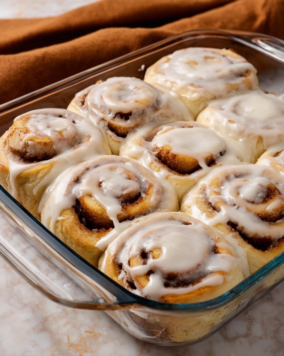 A clear glass baking dish holds seven cinnamon rolls closely packed together. Each roll has a visible swirl pattern with a golden-brown inner layer of cinnamon filling and a light, soft dough base. A thick layer of creamy white icing is generously spread over each roll, partially covering the swirls and dripping slightly into the spaces between them. The dish sits on a white marbled surface with a soft brown cloth in the background. photo taken with an iphone --ar 4:5 --v 7