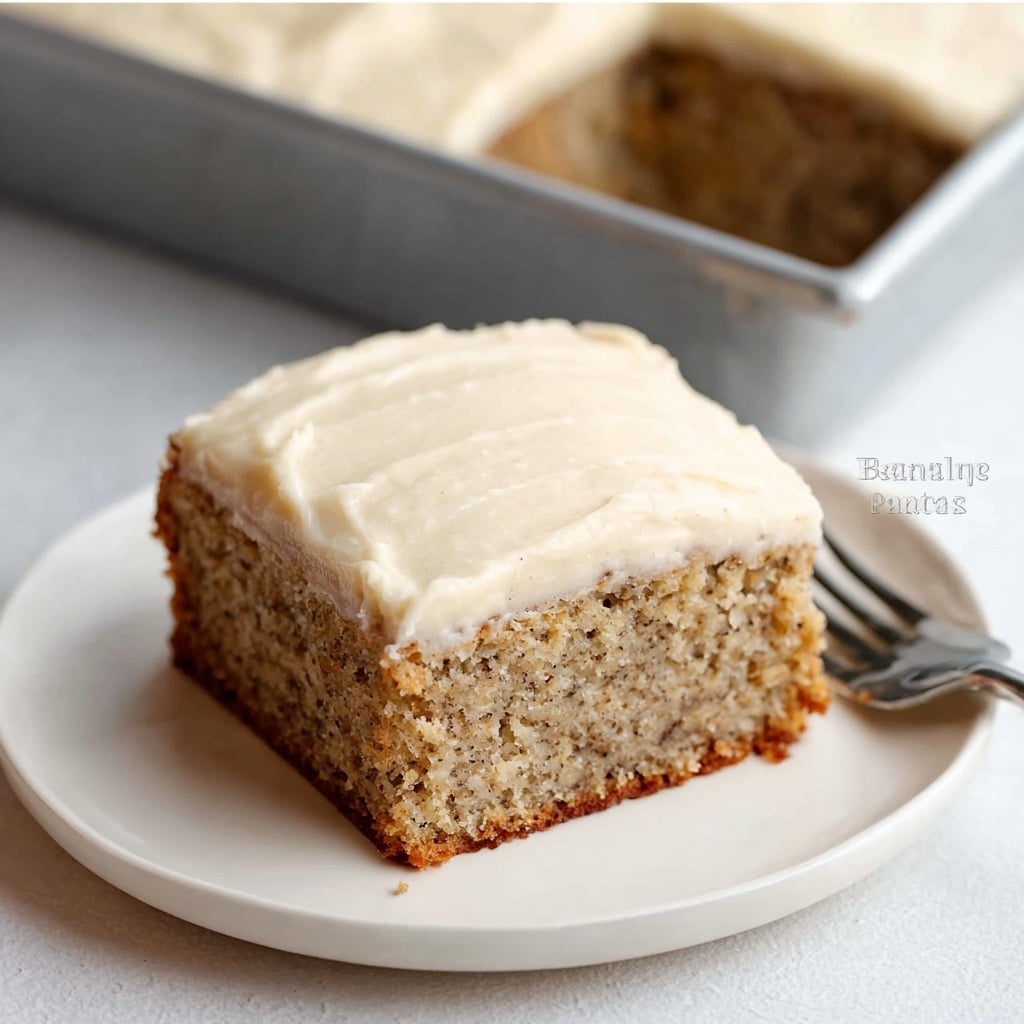 A square slice of banana cake sits on a white round plate with a fork next to it. The cake has one thick, light brown layer with a soft, moist texture speckled with tiny dark spots from the banana. On top, there is a thick, smooth layer of creamy white frosting, spread evenly with slight ridges. Behind the plate, the rest of the rectangular cake with the same frosting layer is visible on a silver tray. The scene is on a white marbled surface. photo taken with an iphone --ar 4:5 --v 7