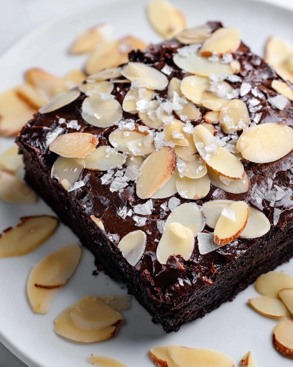 A single square piece of dark chocolate brownie topped with a thick layer of slivered almonds scattered unevenly across the surface, with visible flakes of coarse salt sprinkled on top adding texture and shine. The rich, moist brownie base has a slightly crinkled, matte finish, contrasting with the shiny, pale cream and light brown almond slices. The brownie sits on a white plate, placed on a white marbled surface with some almond slices spread around it loosely. photo taken with an iphone --ar 4:5 --v 7