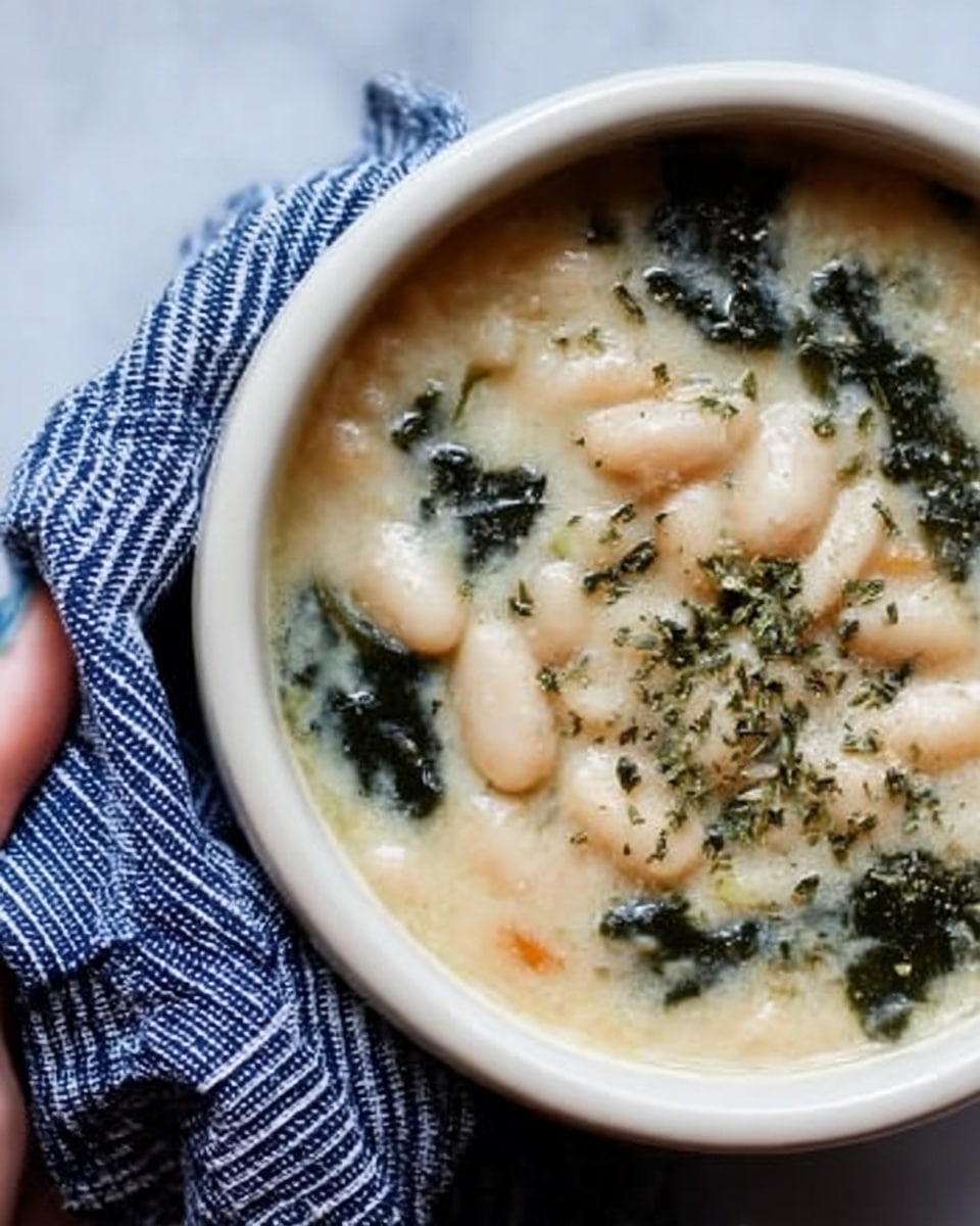The image shows a bowl filled with creamy white bean soup that has a smooth, thick texture. The soup contains soft white beans and pieces of green leafy herbs scattered on top. There is a light sprinkle of dried herbs adding texture and color contrast. The bowl is deep and white, placed on a white marbled surface. Next to the bowl, a woman’s hand is holding a blue and white striped cloth, adding a touch of coziness to the scene. Photo taken with an iphone --ar 4:5 --v 7