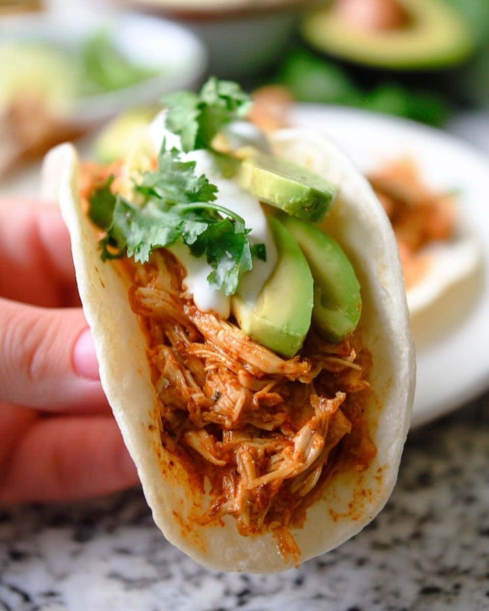 A close-up image of a soft white tortilla taco held by a woman's hand, filled with a thick layer of shredded, orange-brown cooked chicken at the bottom, topped with a dollop of white sour cream, a few slices of bright green avocado, and a small sprig of fresh green cilantro leaves on top. In the blurred background, there is a white plate on a white marbled surface with more food partially visible. Photo taken with an iphone --ar 4:5 --v 7
