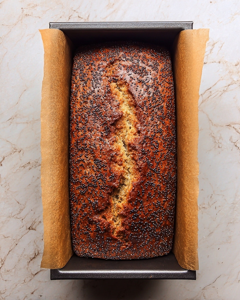 This image shows a freshly baked rectangular loaf of golden brown bread with a cracked top surface covered in small, round dark poppy seeds. The loaf is still inside a dark metal baking tin lined with parchment paper on the longer sides. The bread looks dense and moist with a slightly rough texture on the top. The background is a white marbled texture. photo taken with an iphone --ar 4:5 --v 7
