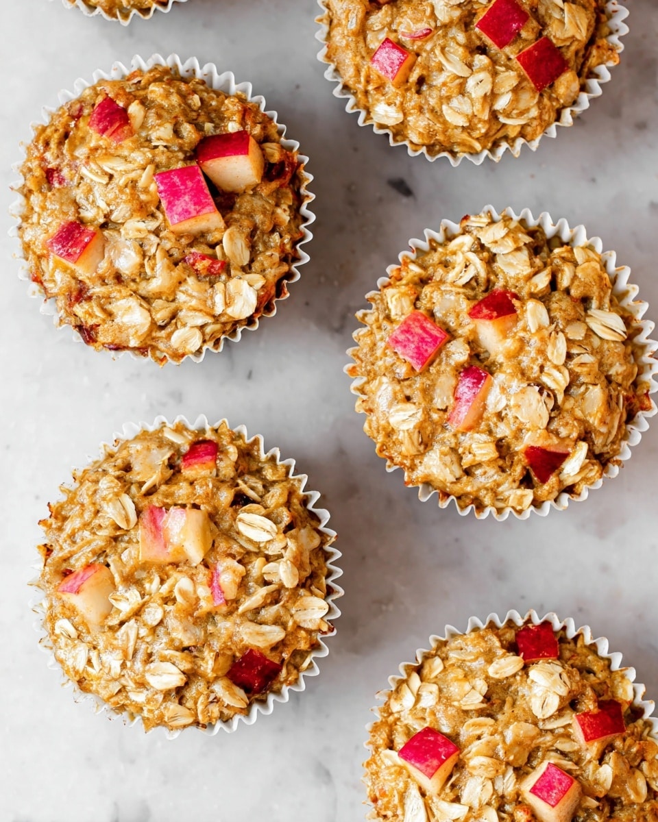 A close-up of five oatmeal muffins with visible oats and small pieces of red apple on top and inside, showing a soft and moist texture with light brown and beige tones mixed with occasional red apple chunks. One muffin in the center is slightly opened with the white paper wrapper peeled down to reveal the dense inside filled with oats and apple pieces. The muffins are placed on a white marbled surface, and blurred whole apples in yellow and red sit in the background. photo taken with an iphone --ar 4:5 --v 7