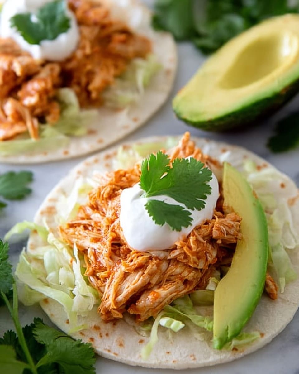 The image shows a close-up of two soft white tortillas on a white marbled surface. On each tortilla, there is a base layer of shredded lettuce, topped with a generous layer of shredded, cooked chicken in a reddish-orange sauce. One tortilla has a dollop of sour cream on top, garnished with a fresh green cilantro leaf. Next to the sour cream, there is a slice of green avocado placed diagonally. In the background, more avocado slices and some scattered cilantro leaves are visible, creating a fresh and colorful presentation. photo taken with an iphone --ar 4:5 --v 7