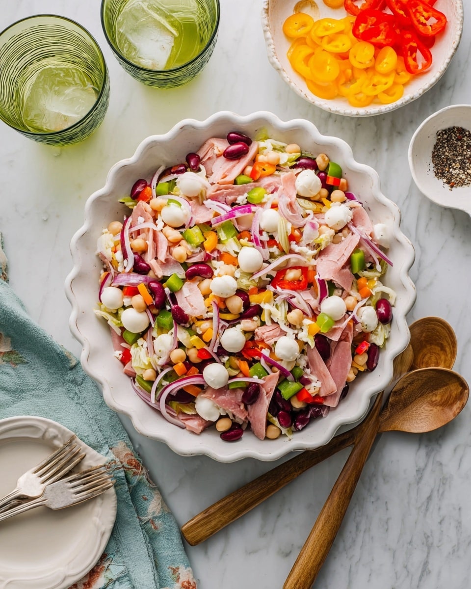 In the white scalloped bowl sits a colorful mixed salad with multiple layers, including pale pink strips of deli meat, bright white small mozzarella balls, dark red kidney beans, light beige chickpeas, and thin slices of red onion. Also sprinkled throughout are pieces of green bell pepper and small bits of orange and yellow peppers, adding vibrant color. Two wooden serving spoons rest on the right side of the bowl. The setting includes a white marbled surface with a small white bowl of sliced yellow and orange peppers above, a white scalloped dish of cracked black pepper to the right, a white napkin on the left, and two greenish glass cups with ice near the top left. Another white plate with silver forks is partially visible at the bottom. photo taken with an iphone --ar 4:5 --v 7