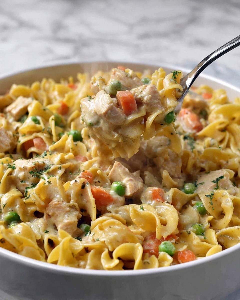 A white bowl filled with creamy chicken noodle casserole features a mix of three main layers: the base layer of wavy, golden-yellow egg noodles with a soft, slightly glossy texture; the middle layer of tender light brown chicken chunks mixed with small green peas and bright orange diced carrots, blending smoothly with the sauce; and the top layer of creamy, beige sauce that coats all ingredients evenly, giving a rich, slightly shiny finish. Steam rises gently from the dish, highlighting its warmth, while a silver fork lifts a scoop of noodles and chicken from the bowl, capturing the texture and mix on the utensil. The whole setup rests on a white marbled surface. photo taken with an iphone --ar 4:5 --v 7