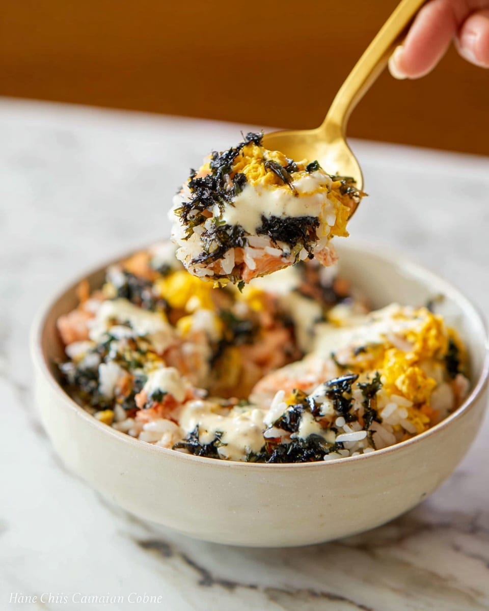 A close-up of a spoonful of a mixed rice dish lifted above a white bowl, showing layers of white rice, black seaweed pieces, bright yellow scrambled eggs, and small bits of light pink fish, all topped with a creamy off-white sauce; the bowl sits on a white marbled surface, and the spoon is a warm golden color held by a woman's hand just out of frame. photo taken with an iphone --ar 4:5 --v 7