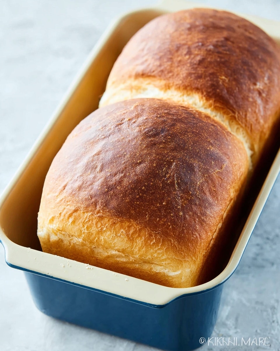 A close-up view of a freshly baked loaf of bread with two rounded, golden brown tops that have a smooth but slightly cracked texture. The bread is inside a blue bread pan with a cream-colored interior, sitting on a white marbled surface. The crust is soft and light brown on one side, darker on the other, showing a perfect bake. Photo taken with an iphone --ar 4:5 --v 7