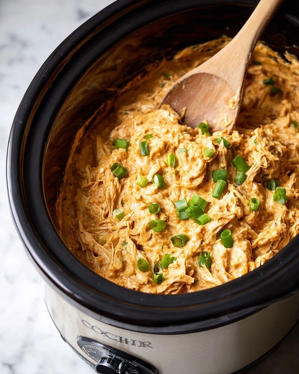 A close-up of a slow cooker filled with a thick, creamy orange dip mixed with shredded white chicken pieces visible throughout. The dip has a rich texture with specks of green herbs mixed in. The top is garnished with bright green sliced scallions scattered evenly. A wooden spoon rests on the side inside the cooker, slightly submerged in the dip. The slow cooker has a silver body with black trim and a dial on the front, sitting on a white marbled surface. Photo taken with an iphone --ar 4:5 --v 7