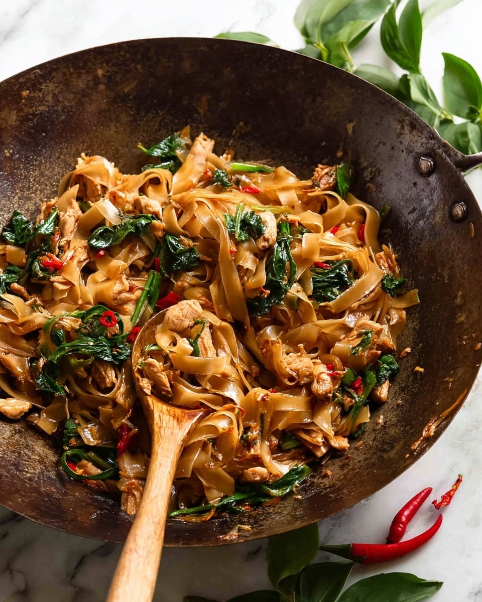 The image shows a rustic dark metal wok filled with glossy stir-fried flat rice noodles that are golden brown, mixed with pieces of tender light brown chicken and bright green leafy vegetables. The noodles are coated with a shiny sauce, and there are small red chili flakes scattered throughout. A wooden spoon is resting inside the wok, partially lifting some noodles, and the wok is placed on a white marbled surface with fresh green leaves and red chili peppers nearby. photo taken with an iphone --ar 4:5 --v 7