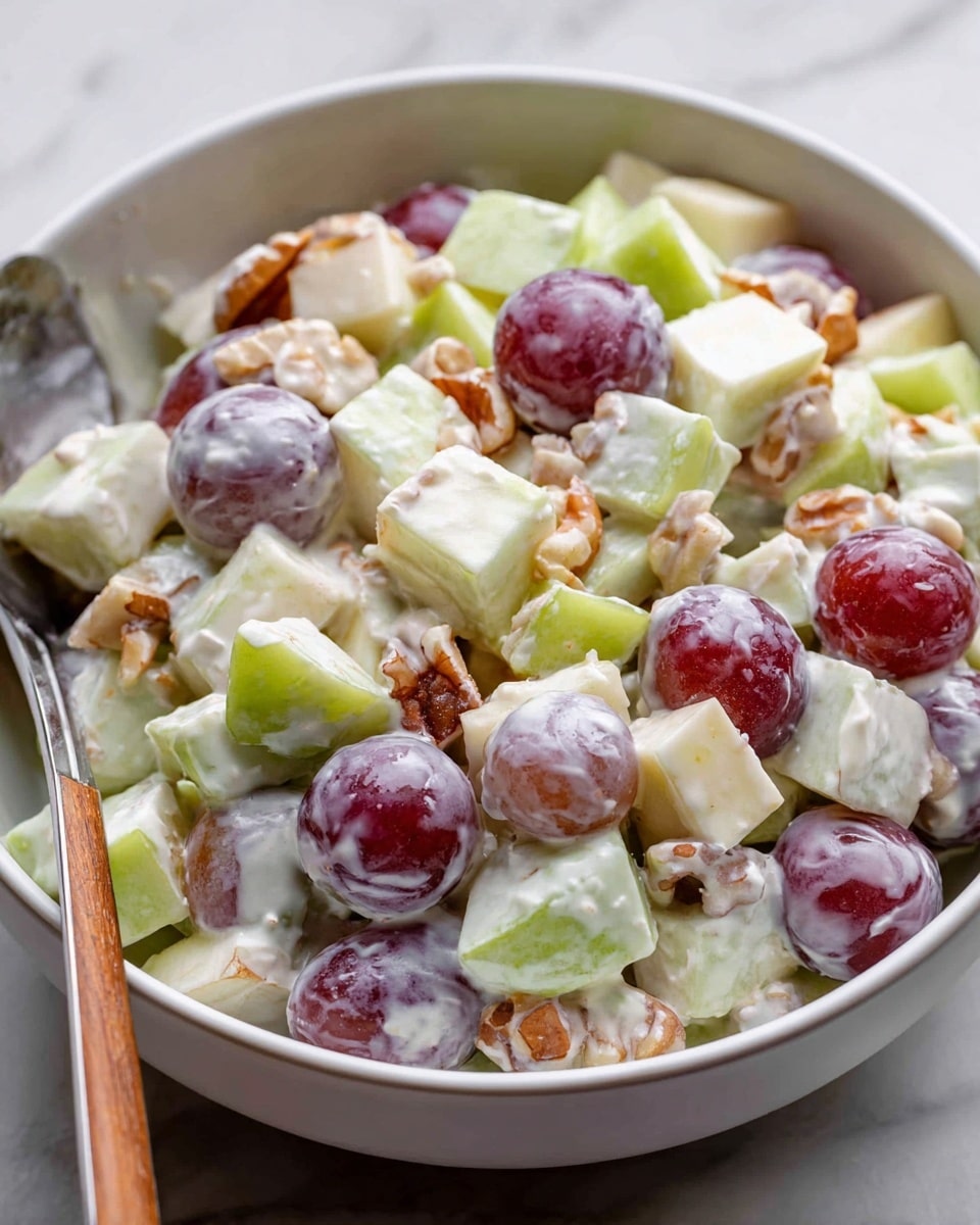 A close-up view of a white bowl filled with a creamy fruit salad. The salad has three main layers: light green apple pieces cut into small cubes, red and purple halved grapes with shiny skins, and chopped light brown nuts mixed evenly throughout. The creamy white dressing coats every ingredient, giving the dish a smooth, thick texture. A metal spoon rests inside the bowl on the left side, partially covered by the salad. The bowl sits on a white marbled surface. photo taken with an iphone --ar 4:5 --v 7