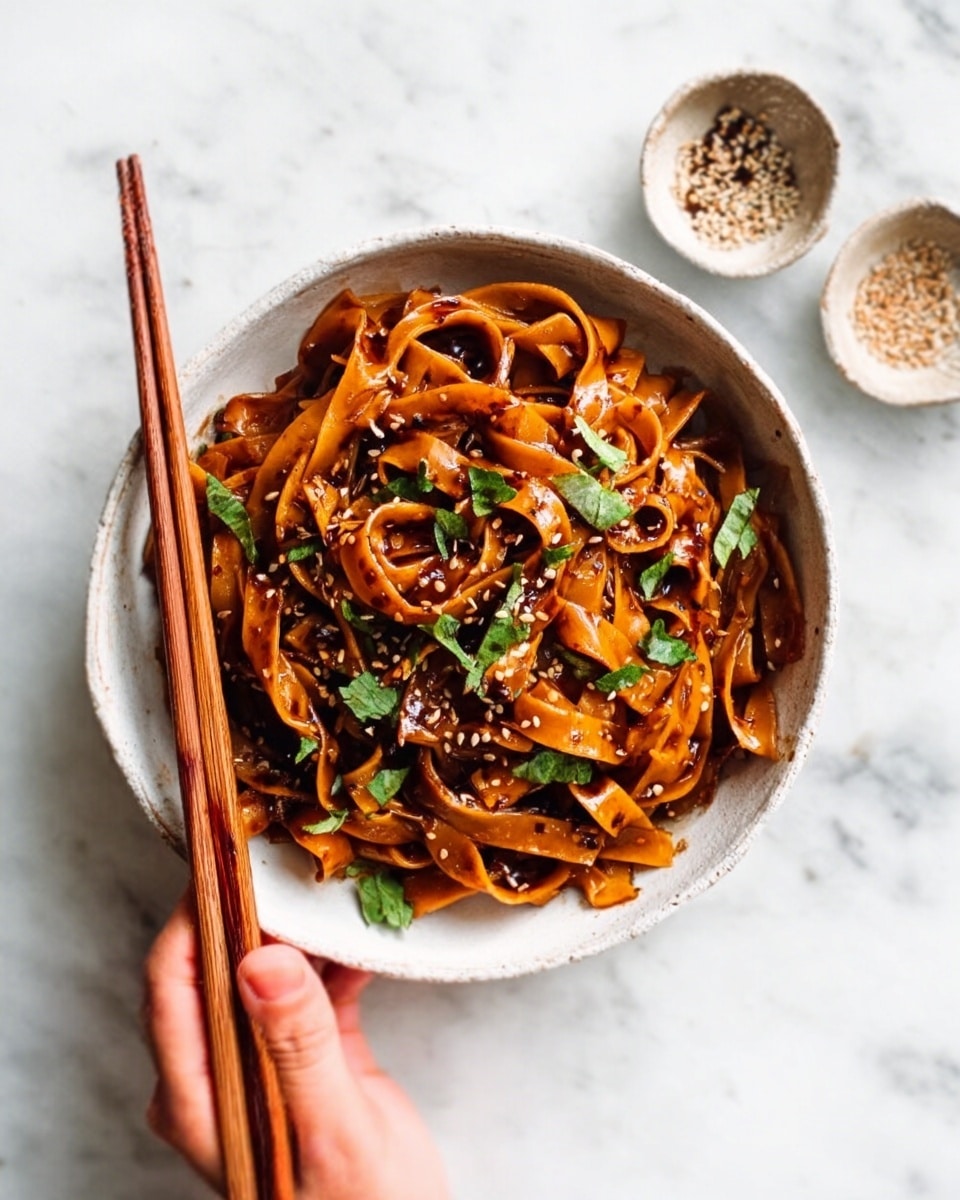 A white bowl filled with thick, flat noodles covered in a shiny, dark reddish-brown sauce. The noodles are twisted and layered in a loose pile. Small green herb pieces and sesame seeds are sprinkled on top, adding contrast. A pair of brown chopsticks rests on the bowl's edge, with a woman's hand gently holding one side. The background features a white marbled surface with small bowls partially visible around the main dish. photo taken with an iphone --ar 4:5 --v 7
