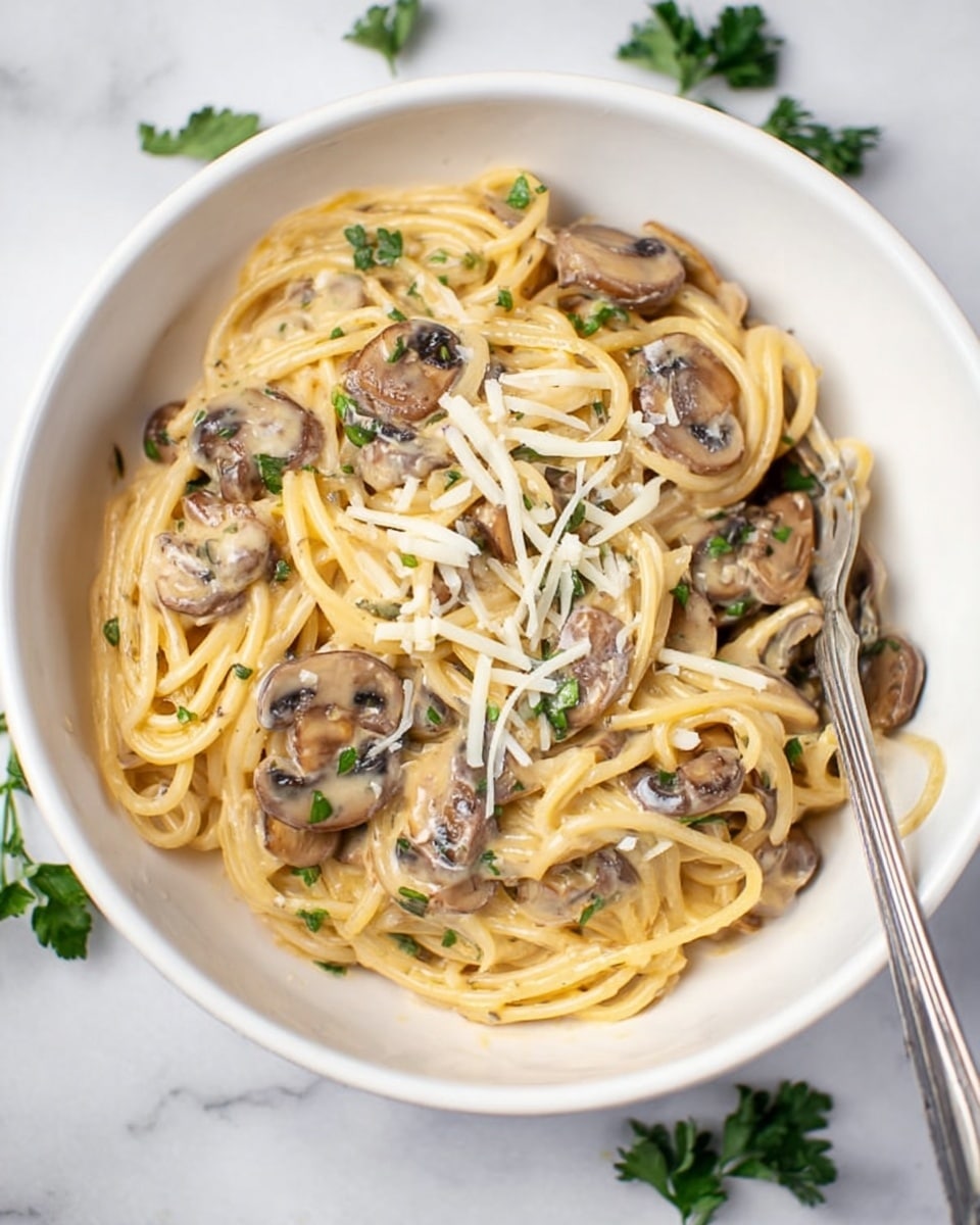 A white bowl holds creamy spaghetti mixed with round, sliced mushrooms that are light brown with darker edges. The pasta is coated in a smooth, pale yellow sauce, with small pieces of green herbs sprinkled throughout. On top, thin shreds of white cheese are scattered over the noodles and mushrooms. The bowl sits on a white marbled surface with a few green herb leaves near it. A silver fork is placed inside the bowl on the right side, touching the pasta. photo taken with an iphone --ar 4:5 --v 7