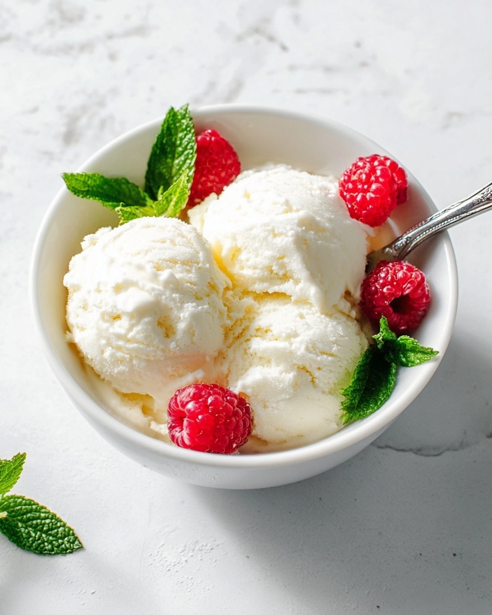A white bowl filled with three scoops of creamy white ice cream, decorated with three bright red raspberries evenly placed around the ice cream. Two fresh green mint leaves sit on top, adding a pop of color and freshness. A silver spoon rests inside the bowl, partly under one scoop. The background shows a white marbled surface with soft natural light enhancing the texture of the ice cream and fruit. photo taken with an iphone --ar 4:5 --v 7