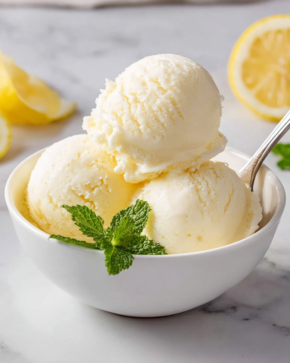 A white bowl holds three smooth, creamy scoops of pale yellow lemon ice cream stacked in a pyramid shape, with the top scoop resting gently on the two below. Behind the scoops, there is a fresh green mint leaf and a bright yellow lemon slice arranged for decoration. A silver spoon is tucked into the right side of the bowl. The bowl sits on a white marbled surface with soft, natural lighting highlighting the icy texture and fresh colors of the dessert and garnishes. Photo taken with an iphone --ar 4:5 --v 7