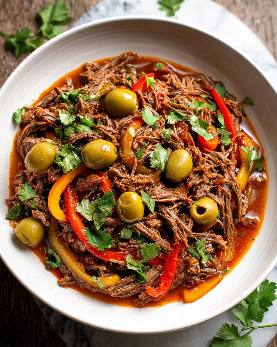 A white bowl is filled with shredded brown beef cooked in a rich reddish-brown sauce, mixed with large green olives and thin strips of red and yellow bell peppers. Fresh green cilantro leaves are scattered on top and around the edges of the dish, adding a touch of bright green color. The bowl is placed on a white marbled surface, highlighting the warm colors of the food inside. Photo taken with an iphone --ar 4:5 --v 7