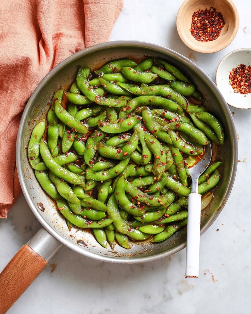 A shiny silver pan filled with bright green edamame pods coated in a glossy sauce with red chili flakes scattered on top. The edamame pods form one thick layer covering most of the pan’s bottom, with a silver spoon with a white handle resting on the right side, slightly under some pods. To the top right, a small white bowl contains extra red chili flakes, and a pale wooden container is partially visible. On the left side, a peach-colored cloth is draped, all placed on a white marbled surface. photo taken with an iphone --ar 4:5 --v 7