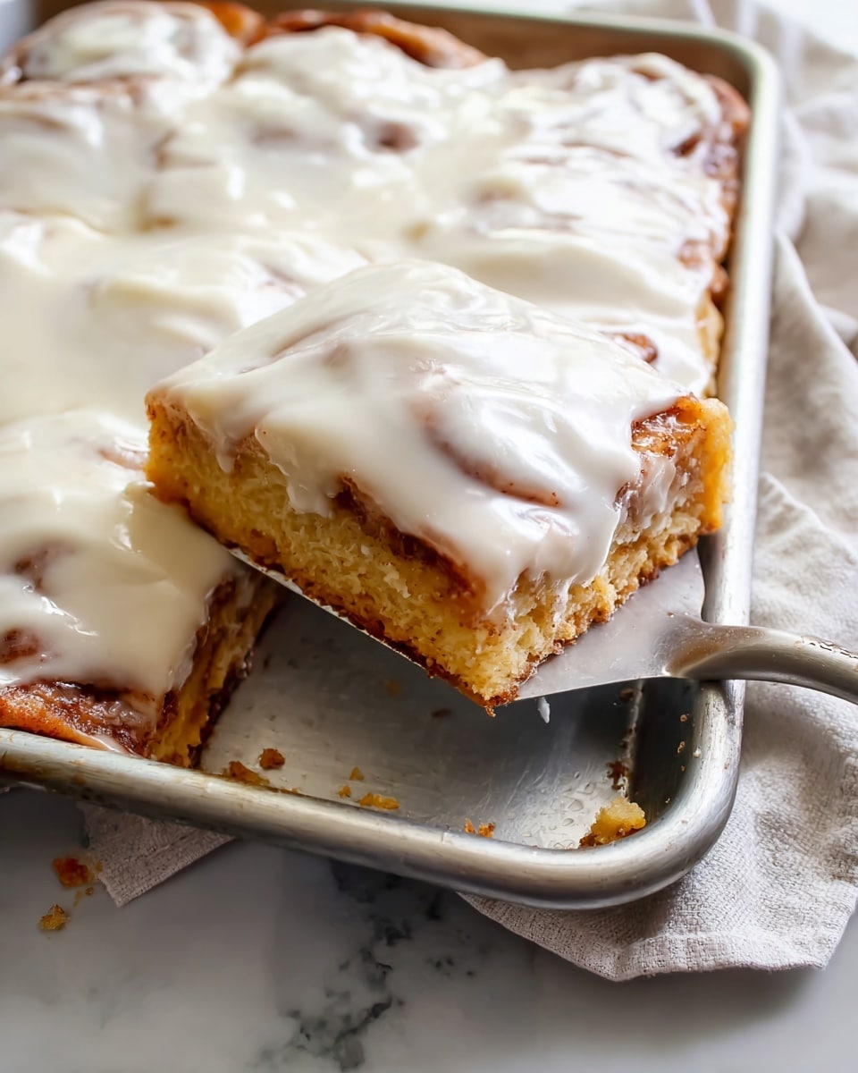 A square piece of cinnamon roll biscuit with creamy white icing is being lifted by a spatula from a silver baking tray. The cinnamon roll biscuit has a light golden brown base layer, topped with swirls of cinnamon filling visible through a smooth, glossy white icing layer covering the entire surface. The edges of the tray show some baked cinnamon filling spilling slightly over the sides. The tray sits on a white marbled surface with a soft cloth nearby, enhancing the cozy, fresh-out-of-the-oven feel. photo taken with an iphone --ar 4:5 --v 7