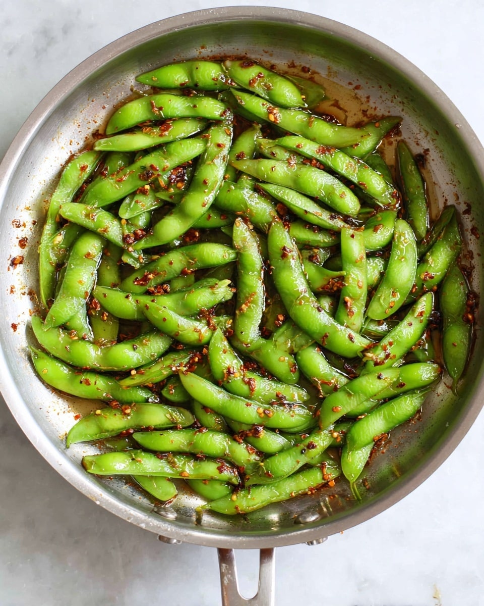 A shiny silver pan is filled with bright green edamame pods cooked in a thin layer of oil and small bits of dark red chili flakes and seasoning scattered evenly on and under the pods, giving a glossy and slightly spicy appearance. The pods are roughly spread in a single layer inside the pan, which sits on a white marbled texture. photo taken with an iphone --ar 4:5 --v 7