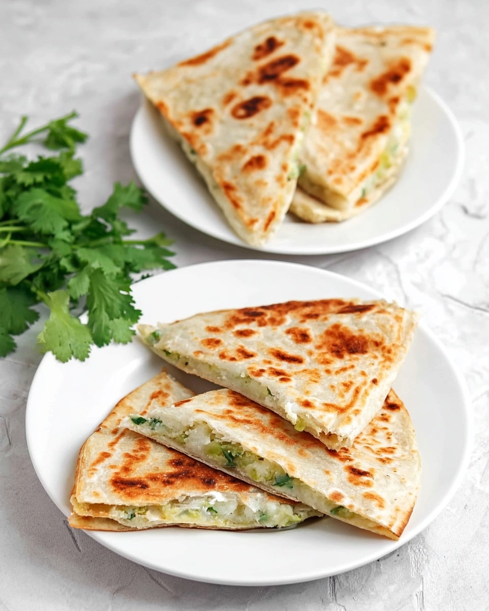 The image shows two white plates on a white marbled surface. The plate in the front holds four triangular pieces of a stuffed flatbread, each piece golden brown with darker brown cooked spots. The inside filling looks soft and light green, suggesting a vegetable mix, and the edges of the flatbread are slightly crispy. Behind it, a second white plate holds two larger folded pieces of the same flatbread, showing the same golden brown and spotted texture. On the left side of the image, some fresh green cilantro leaves are placed on the white marbled surface. photo taken with an iphone --ar 4:5 --v 7
