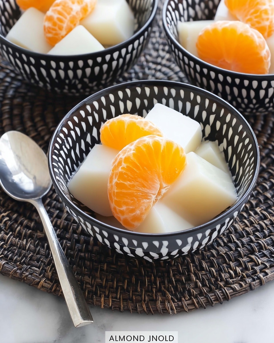 The image shows a small black bowl with white dash patterns filled with white almond jello cut into smooth, translucent cubes. Among the jello cubes are bright orange mandarin orange slices with a shiny, juicy texture. The bowl sits on a woven dark placemat, and a silver spoon lies beside it on the left. Two more bowls with the same pattern and contents are partially visible in the background. The whole scene is set on a white marbled surface. photo taken with an iphone --ar 4:5 --v 7