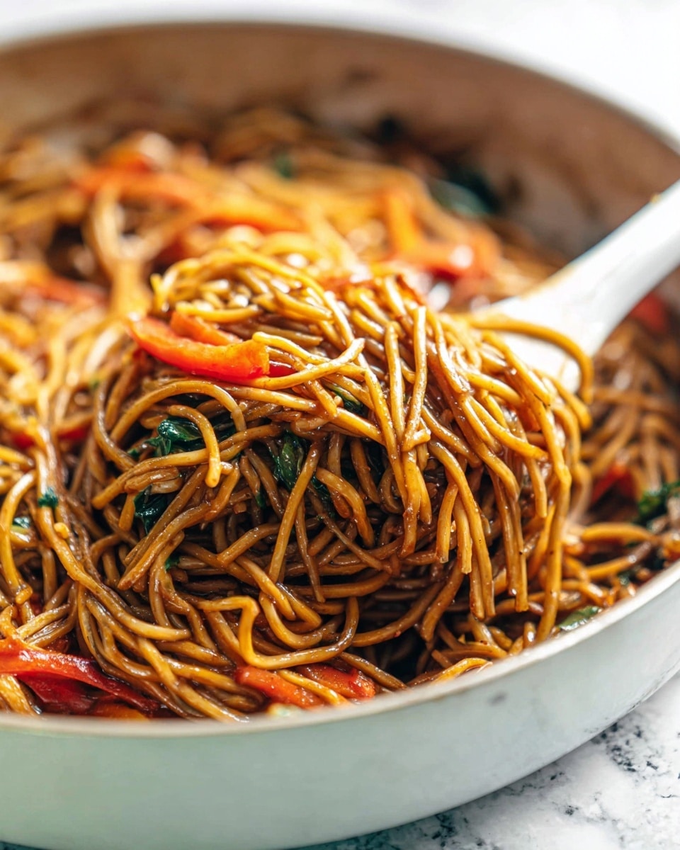 A close-up image of stir-fried noodles piled high over a white frying pan, with glossy, thin strands of brown noodles tangled together. Mixed within the noodles are thin slices of bright orange carrot sticks and small pieces of red bell pepper adding pops of color. A white tongs is lifting a thick bundle of the noodles, showing their shiny and slightly oily texture with some green leafy bits scattered throughout. The background is a soft white marbled texture, giving a clean and fresh look to the scene. photo taken with an iphone --ar 4:5 --v 7