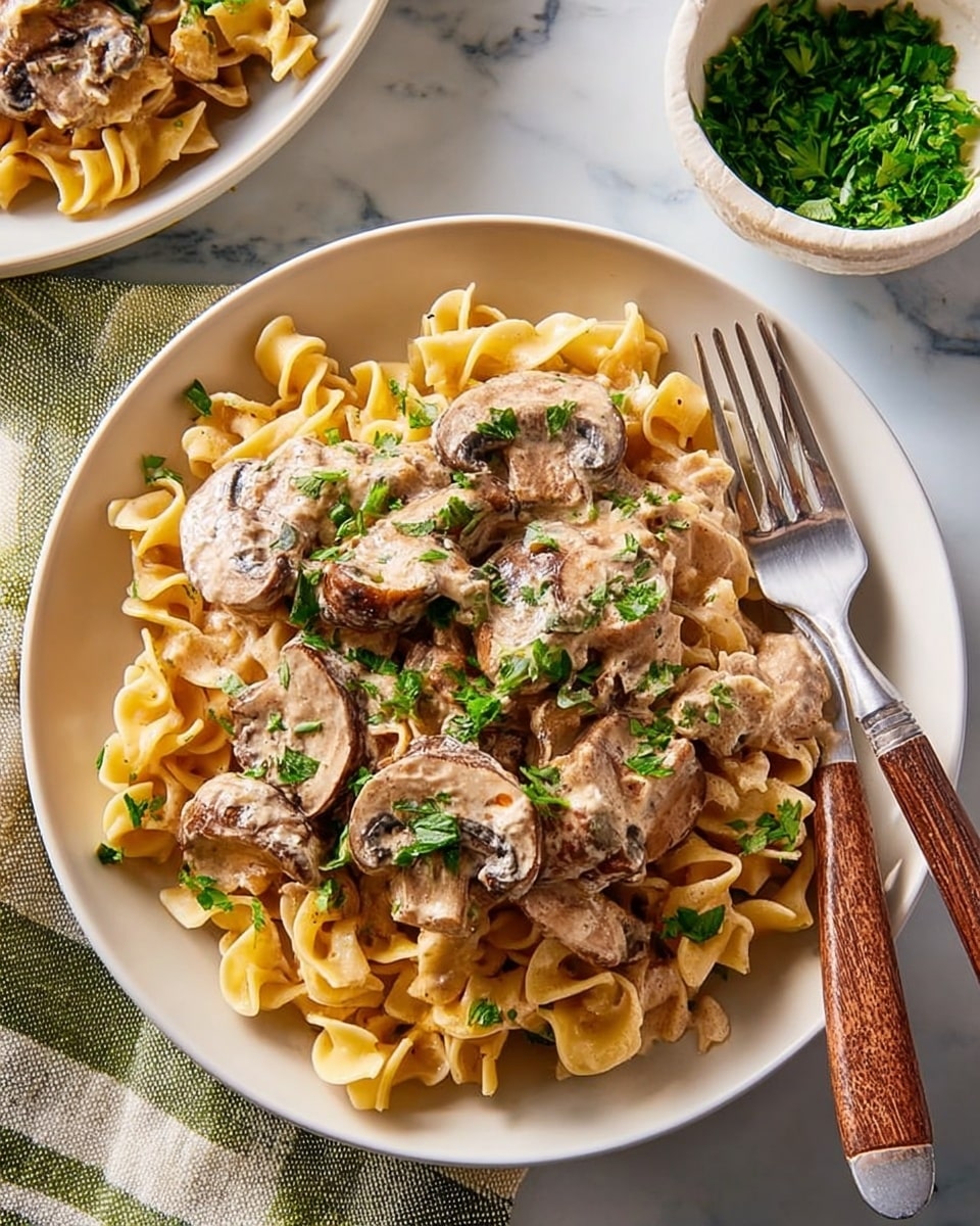 A white plate is filled with three layers: at the bottom, pale yellow egg noodles with a curly texture spread out evenly; on top, a creamy light brown sauce generously covering the noodles; and layered over that, thick slices of cooked mushrooms with a soft, slightly shiny texture. Small green parsley leaves are sprinkled across the whole dish adding bright green spots. A fork with a light wooden handle and silver prongs rests on the right side of the plate. The plate sits on a white marbled surface with a white and green striped cloth napkin next to it. In the background, a small white bowl with chopped green herbs is visible. Photo taken with an iphone --ar 4:5 --v 7