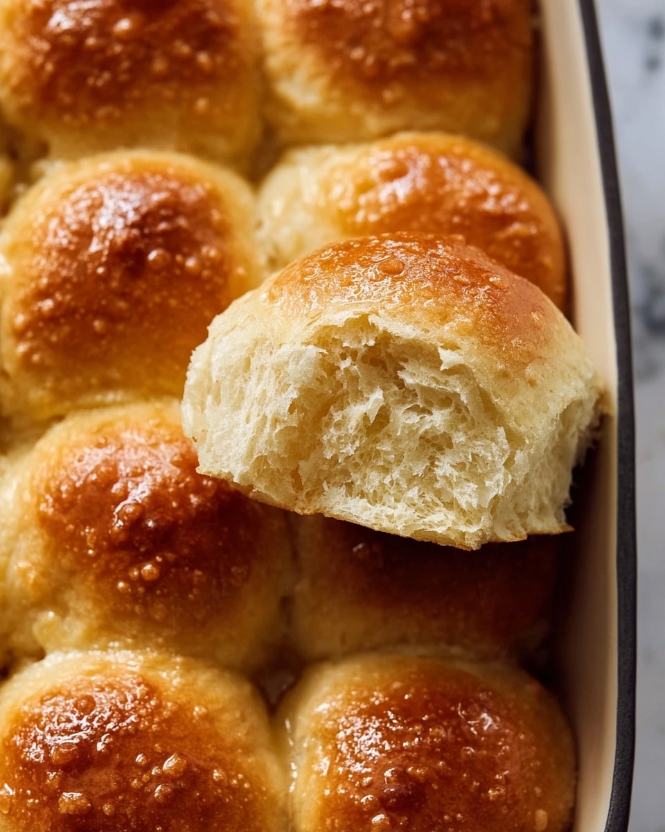 A white scalloped-edge ceramic dish filled with nine golden brown dinner rolls, each roll showing a slightly glossy, smooth, and soft texture with subtle browning on the rounded tops. The rolls are closely packed, rising above the dish’s edges, with a light and airy appearance. The dish rests on a white marbled surface. Photo taken with an iphone --ar 4:5 --v 7