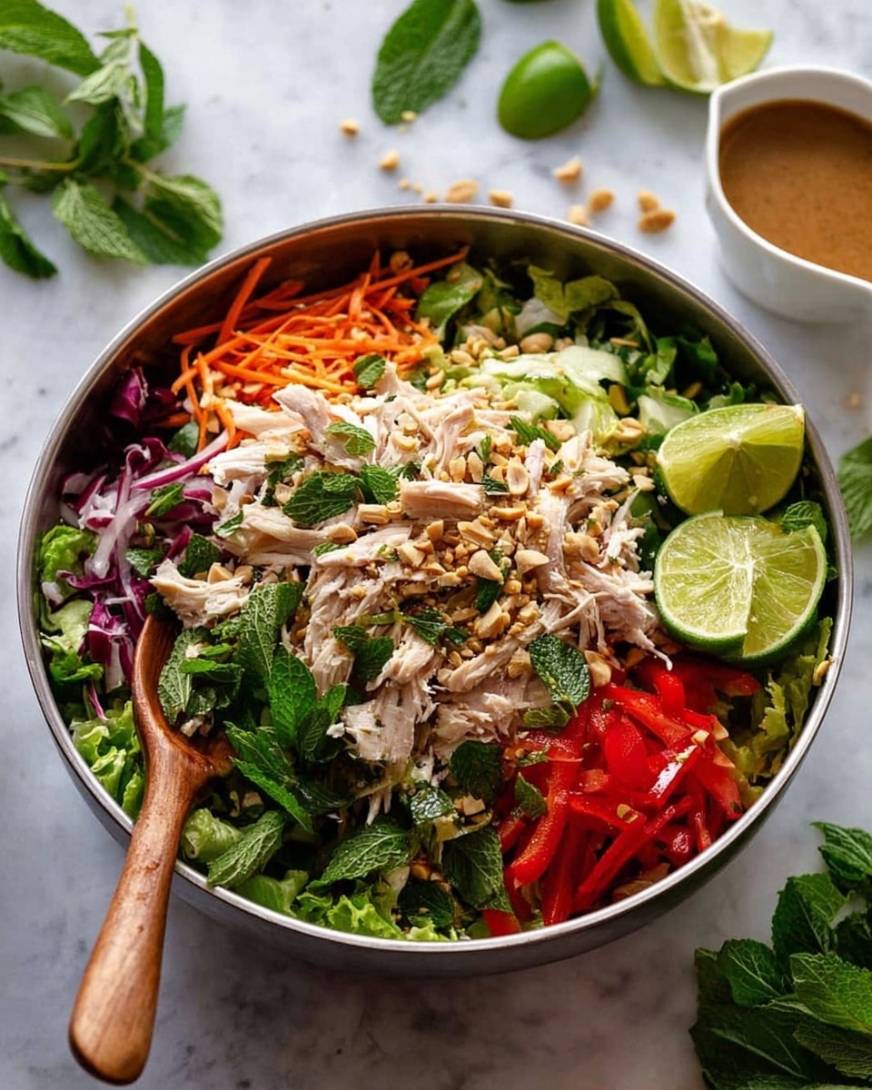 A large silver bowl filled with a colorful salad that has several layers: the bottom layer is chopped green lettuce, above it are shredded white chicken pieces, thin slices of purple onion, red bell pepper strips, and orange carrot shreds, topped with crushed peanuts and fresh green herbs like cilantro and mint leaves. On the right side of the salad, there are three lime wedges placed on top. A wooden spoon and fork rest inside the bowl on the left and right sides. In the background, a small white sauce boat with brown dressing sits, all on a white marbled surface with a few scattered green leaves and lime wedges nearby. Photo taken with an iphone --ar 4:5 --v 7