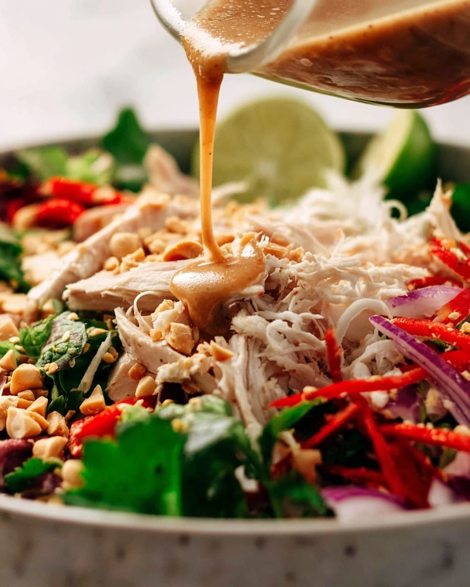 A close-up of a colorful salad in a white bowl with a white marbled background, showing a woman's hand pouring light brown dressing over it. The salad has several layers: at the base, bright green leafy herbs and vegetables; on top of that, white shredded chicken pieces mixed with thin red strips, possibly red bell pepper or chili; scattered crushed light brown nuts spread evenly around; a few slices of red onion and lime wedges in the background add subtle pops of purple and light green. The dressing pours in a smooth stream over the shredded chicken and nuts, giving a fresh and tasty look. Photo taken with an iphone --ar 4:5 --v 7