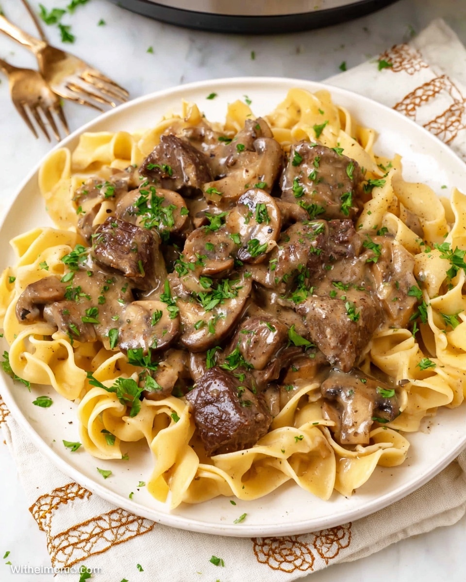 A white plate holds a dish with three main layers: at the bottom there are wide, flat egg noodles that are pale yellow and slightly curled. On top of the noodles are chunks of dark brown cooked beef and sliced mushrooms that are light brown and tender-looking. The whole dish is covered in a glossy, creamy beige sauce dotted with specks of black pepper, garnished with small, fresh green parsley pieces scattered evenly across the top. The plate rests on a white cloth with beige embroidery, all set against a white marbled texture surface in the background. Photo taken with an iphone --ar 4:5 --v 7