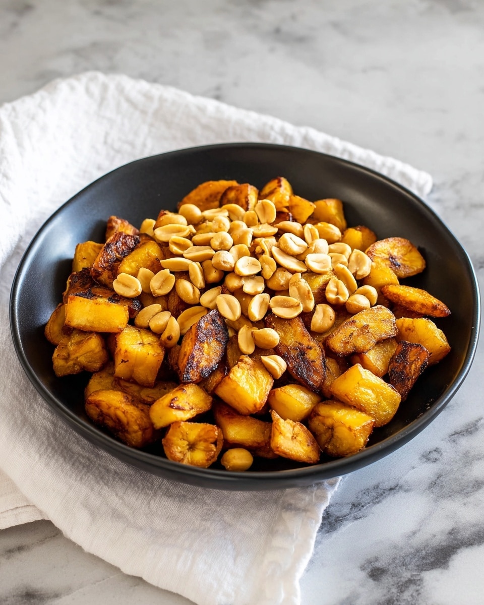 A black bowl full of golden brown fried plantain cubes as the bottom layer, showing a crispy texture with some darker caramelized edges. On top, there is a generous layer of light tan roasted peanuts scattered unevenly across the plantain cubes. The bowl sits on a white marbled surface, with a white towel casually folded and placed in the background. photo taken with an iphone --ar 4:5 --v 7