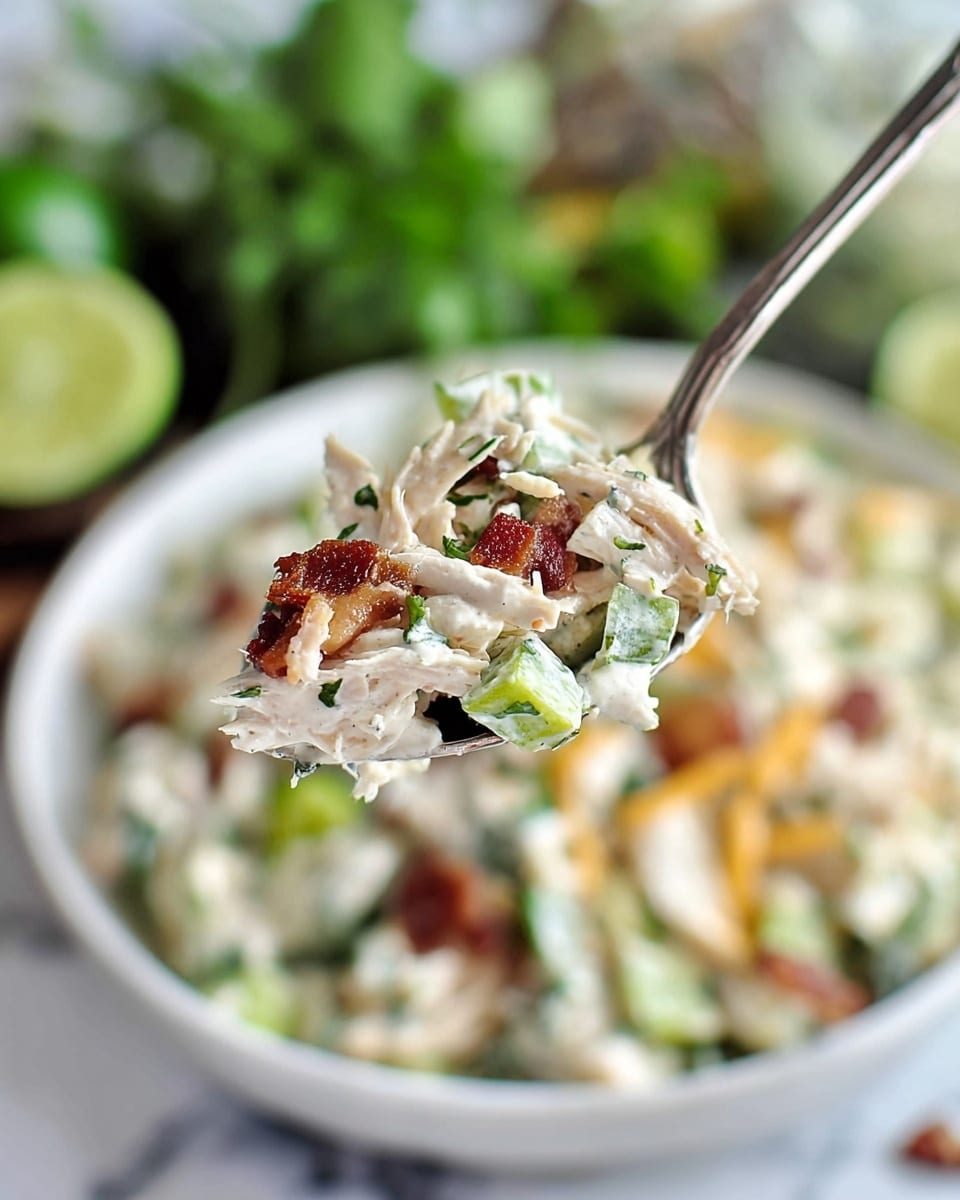 A close-up of a spoon holding a mixed salad with visible layers including shredded light beige chicken, small green celery pieces, white creamy dressing, and scattered reddish-brown bacon bits, with some strands of pale orange cheese; in the background, a white bowl filled with the same salad is slightly blurred on a white marbled surface, with some green herbs and lime wedges also faintly visible. photo taken with an iphone --ar 4:5 --v 7