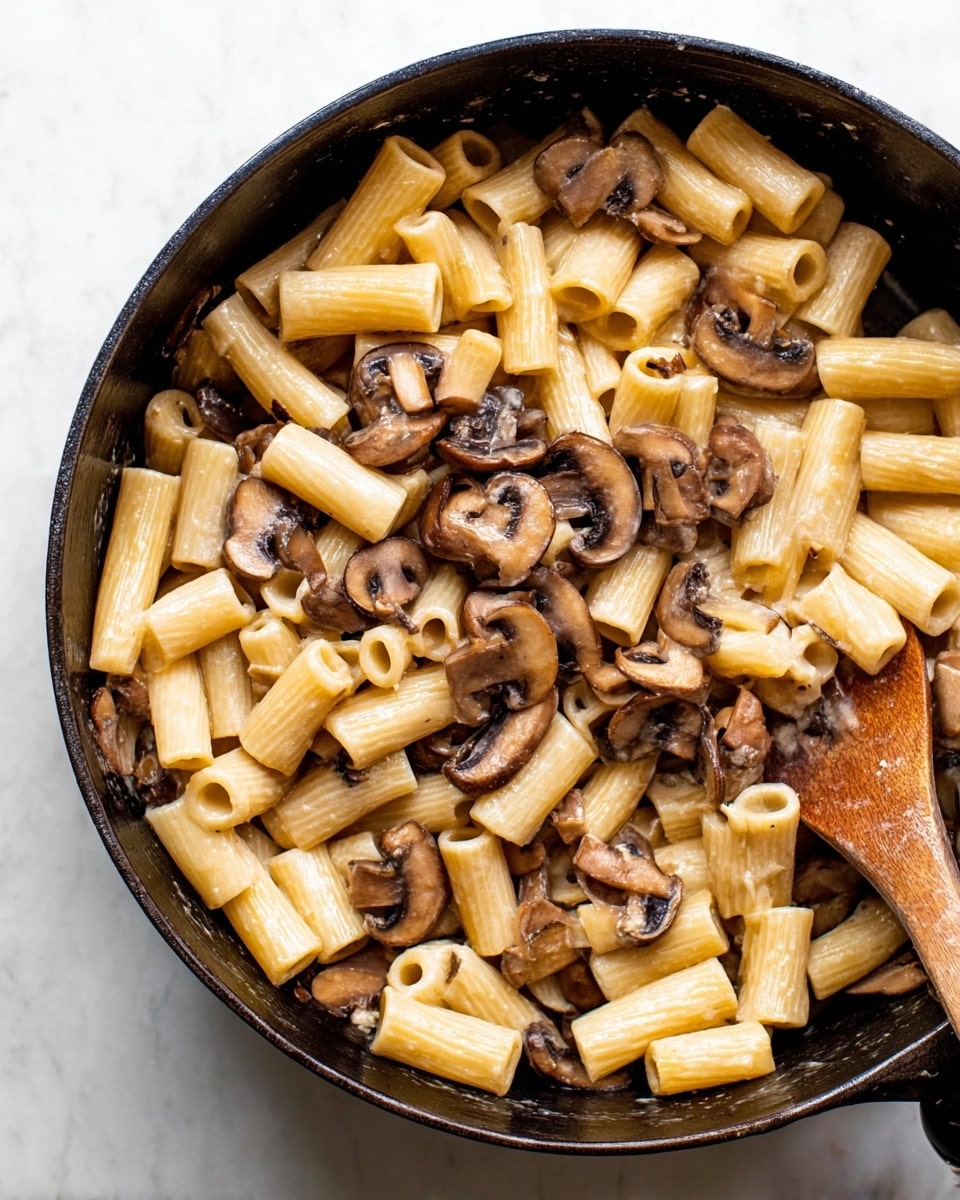 The image shows a black cast-iron pan filled with a creamy pasta dish that has two main layers: a bottom layer of short tube-shaped rigatoni pasta in light beige, smooth and slightly shiny, and a top layer of sliced brown mushrooms with a moist, cooked texture, evenly mixed throughout the pasta. A wooden spoon is partially visible on the right side of the pan, resting inside the dish. The pan sits on a white marbled surface, adding a clean and simple background to the close-up shot. photo taken with an iphone --ar 4:5 --v 7
