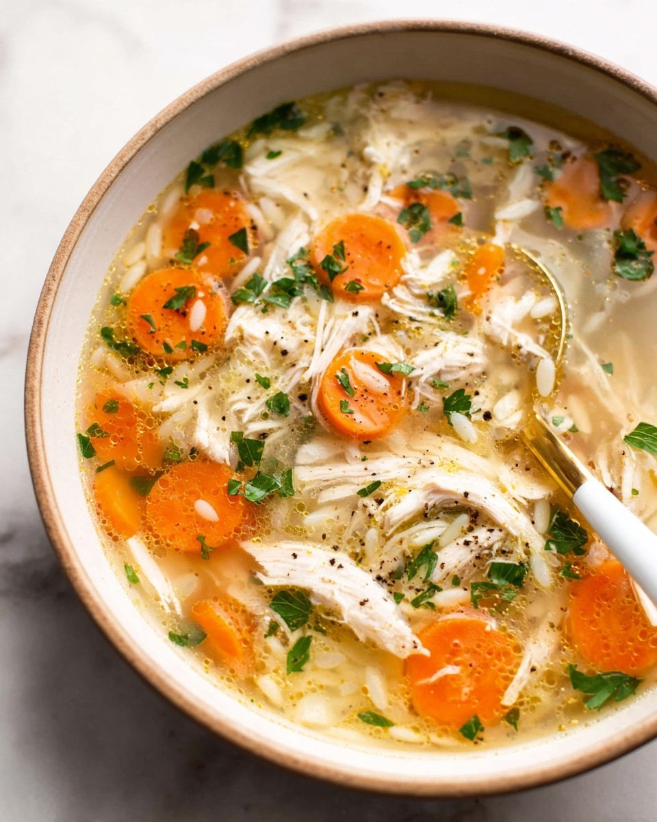 A close-up view of a bowl of chicken soup with clear broth showing layers of shredded white chicken meat scattered across the surface, bright orange carrot slices floating evenly, and small white orzo pasta pieces mixed throughout. Bits of green parsley garnish are sprinkled on top, adding a fresh touch, while black pepper flakes are also visible. The soup is contained in a white bowl with a light brown rim, sitting on a white marbled surface. A spoon with a white handle and golden scoop rests inside the bowl, half-submerged in the broth. Photo taken with an iphone --ar 4:5 --v 7