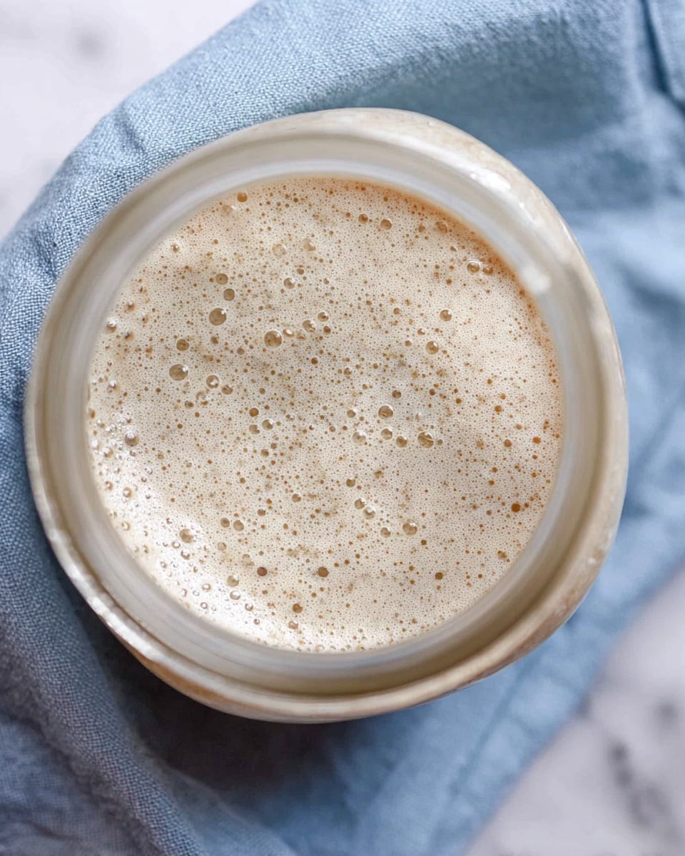 This is a top view of a glass jar filled with a bubbly, light beige mixture that has a frothy texture with small and medium-sized bubbles spread across its surface. The jar sits on a slightly wrinkled blue cloth, which rests on a white marbled texture. The jar's mouth is round, clear, and prominent in the foreground, focusing mainly on the creamy, bubbly mixture inside. photo taken with an iphone --ar 4:5 --v 7