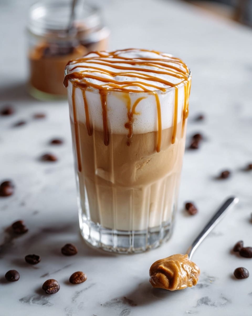 A clear glass filled with three visible layers: a bottom beige coffee layer with swirling light cream mixing in, a middle frothy white foam layer, and a top layer decorated with thick caramel-colored syrup drizzled in wavy lines that slightly drip down the side; the glass sits on a white marbled surface with scattered dark brown coffee beans and a metal spoon nearby holding a dollop of caramel-colored spread, with a blurred background. photo taken with an iphone --ar 4:5 --v 7