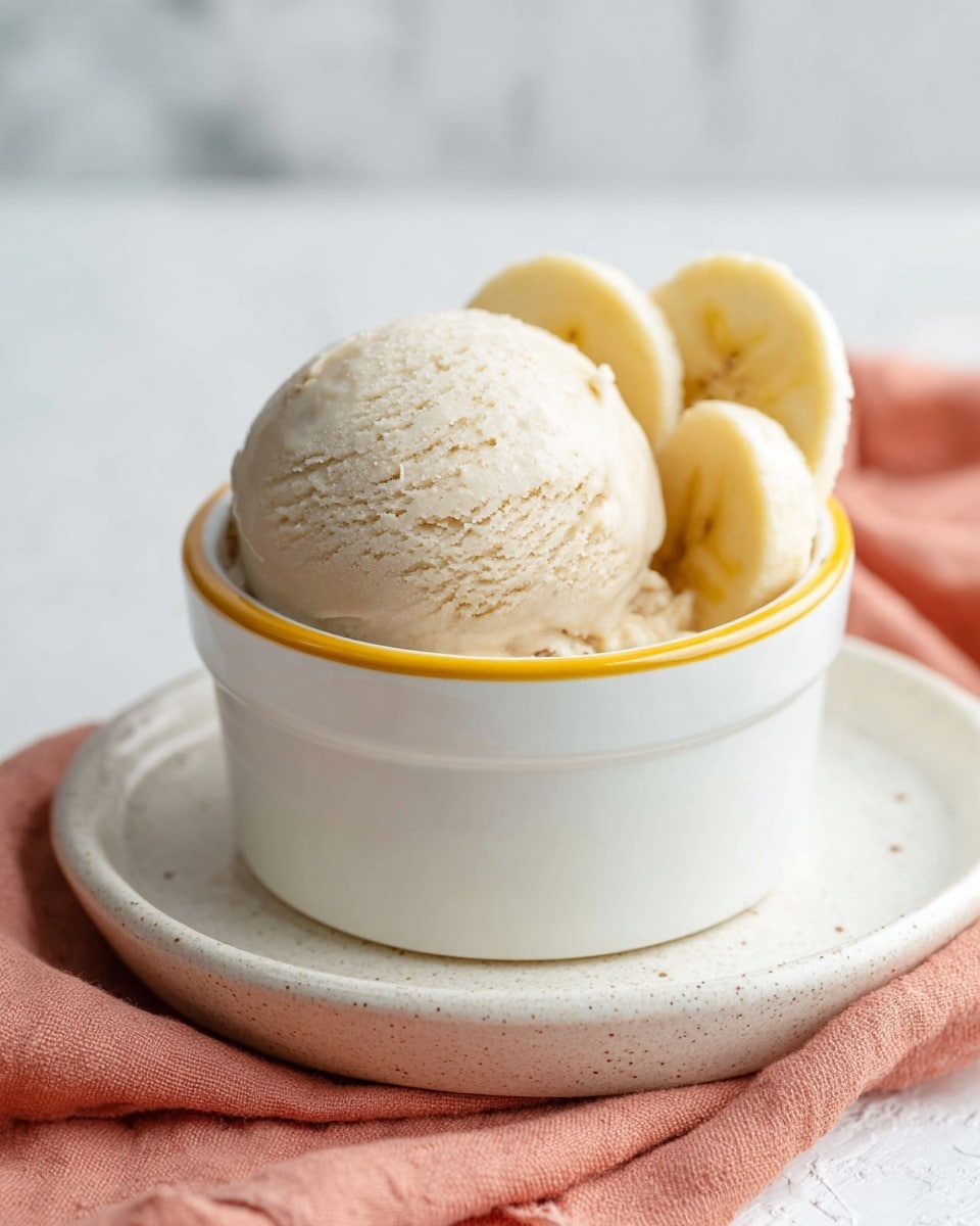 A single scoop of light beige ice cream with a smooth and slightly textured surface sits in a white ramekin with a yellow rim, accompanied by several thick pale yellow banana slices tucked neatly beside the ice cream inside the ramekin. The ramekin is placed on a white plate with a speckled edge, which rests on a soft peach-colored cloth. The background is a white marbled texture, creating a clean and bright setting. photo taken with an iphone --ar 4:5 --v 7