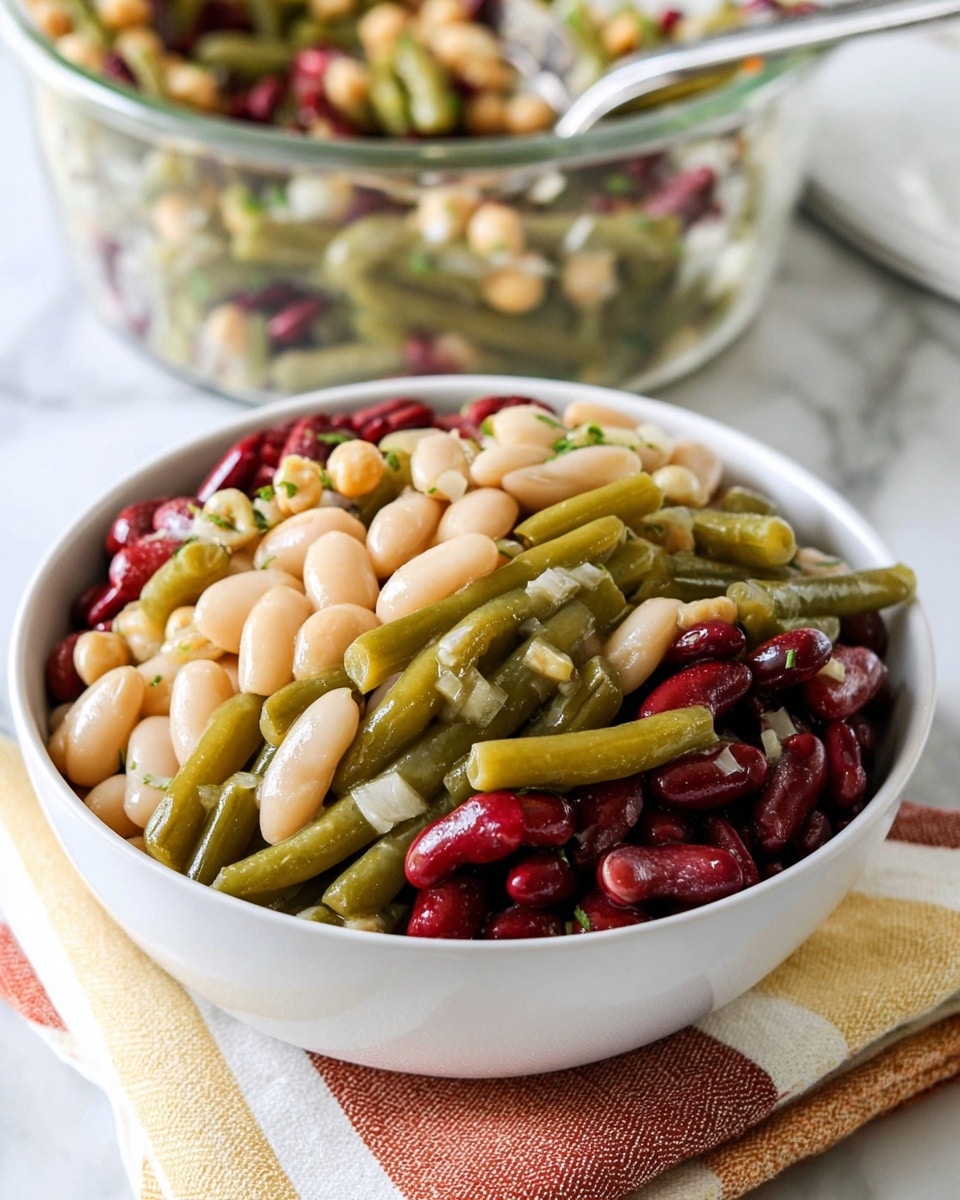 A white bowl filled with a colorful three-layered bean salad sits atop neatly folded striped cloths in shades of cream, yellow, and rust. The bottom layer shows pale, creamy white beans with a smooth texture, mixed closely with pale green beans that have a slight sheen. The middle layer is made of bright green beans that look firm but tender, while the top layer consists of shiny, dark red kidney beans creating a rich contrast. Small bits of chopped herbs and chickpeas are scattered throughout, adding touches of green and light tan. In the background, a clear glass bowl with more of the mixed salad adds depth, set against a white marbled surface. A silver spoon peeks out from the glass bowl. Photo taken with an iphone --ar 4:5 --v 7