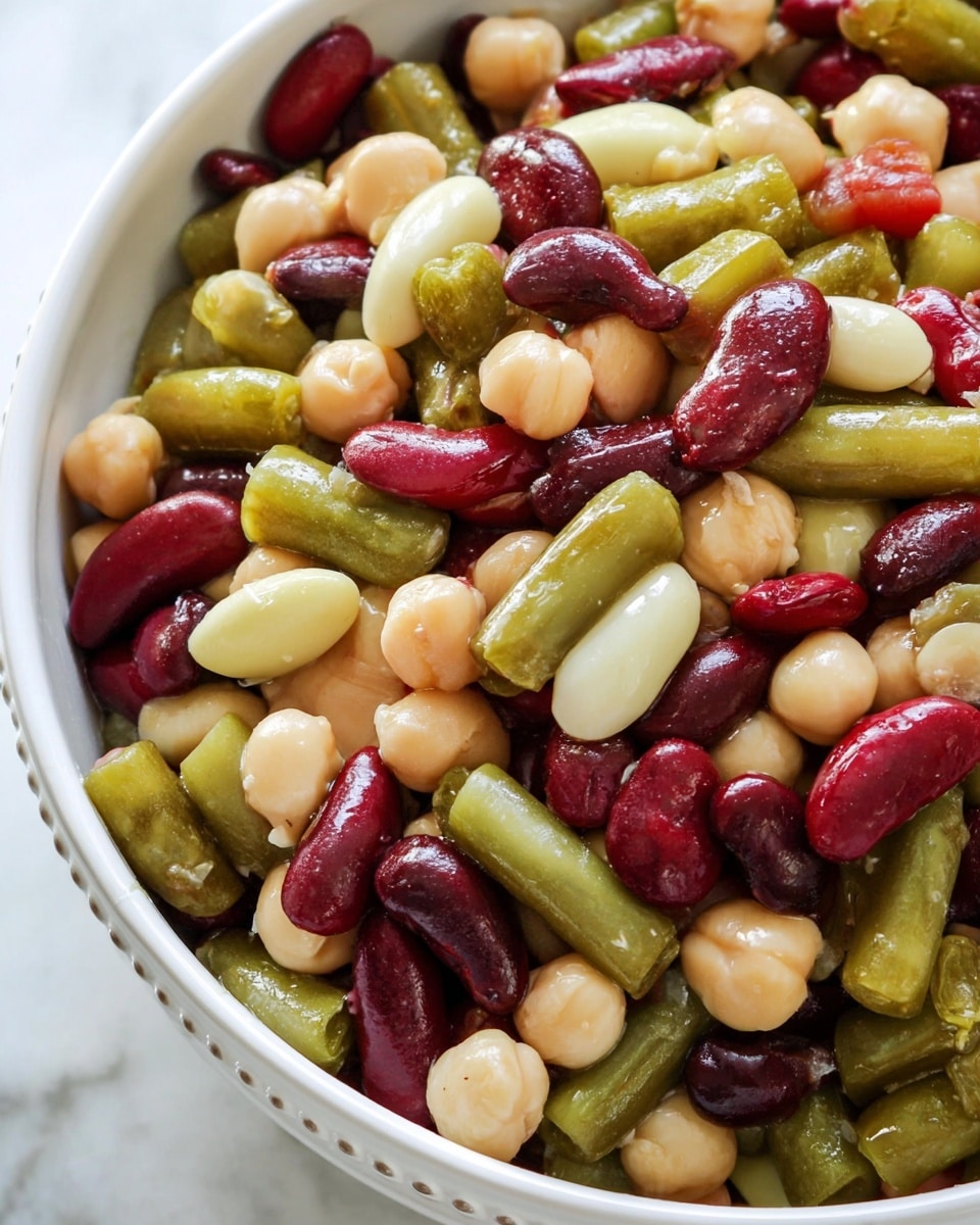 A close-up view of a mixed bean salad in a white bowl, showing multiple layers of colorful beans such as dark red kidney beans, pale green lima beans, and light beige chickpeas, all glistening with a light dressing. The beans are whole and arranged randomly, creating a textured mix of smooth, curved surfaces and soft natural shapes in red, green, and cream shades. The white bowl has a subtle dotted pattern along its rim and the background is a white marbled texture. photo taken with an iphone --ar 4:5 --v 7
